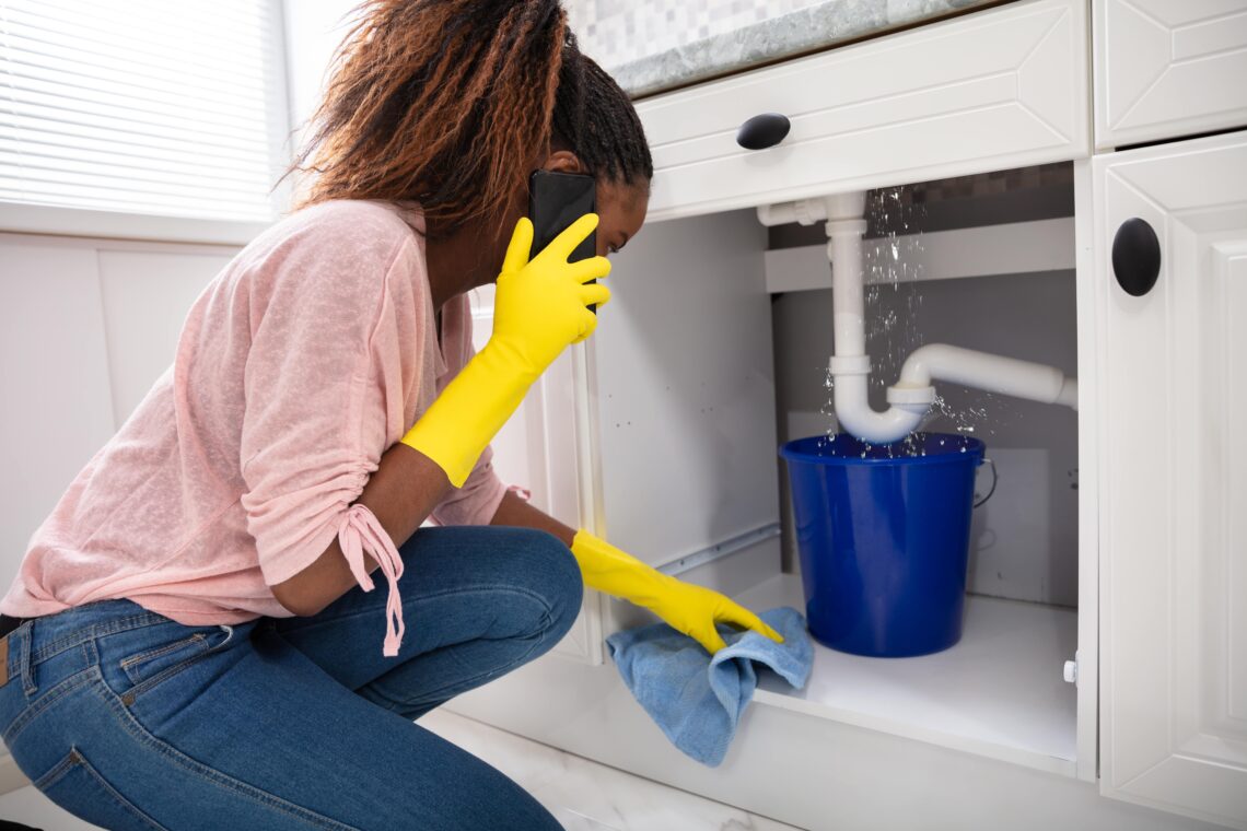 Black Woman Stopping Leak Under Kitchen Sink with Bucket and Towel